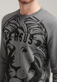 Sweat-shirt gris avec un motif de lion en noir, col de style sweat-shirt et manches longues, fabriqué en tissu doux et texturé.