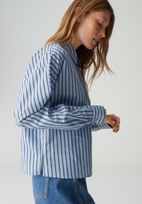 Chemise à manches longues rayée bleu et blanc avec col, présentant une coupe décontractée, des poignets à boutons et des fentes latérales à l'ourlet.