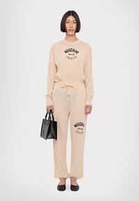 Svijetlosmeđi komplet koji se sastoji od dugog rukava i trenirki s crnim grafikom "Moschino Jeans". Model drži malu crnu torbicu.