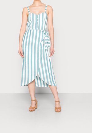 Femme portant une robe midi sans manches à rayures verticales bleues et blanches, boutonnée, avec un nœud sur le côté et des sandales à talons hauts camel, debout sur un fond uni.