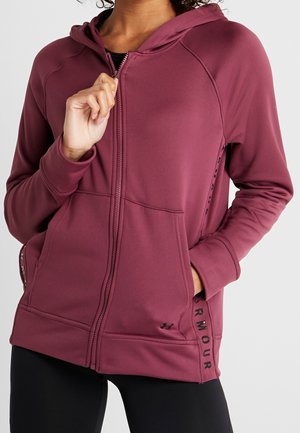 Person trägt einen kastanienbraunen Kapuzenpullover mit Reißverschluss und Seitentaschen sowie schwarze Leggings, Hand hält den Reißverschluss in Brusthöhe.