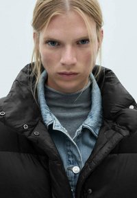 Sort pufferjakke med høj krave, lagdelt over en grå turtleneck og denimskjorte. Har sølvtrykknapper og en glat, skinnende tekstur.