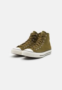 High-top sneakers in olive green canvas with white rubber toe cap and sole, featuring black stripe accents and metal eyelets for laces.