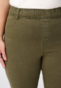 Olijfgroene stretchy broek met een elastische tailleband, voorkruiszakken en een gladde textuur. Beschikt over decoratieve stiksels langs de naden.