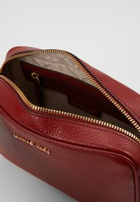 Czerwony skórzany worek Michael Kors z widoczną fakturą i złotym zamkiem, częściowo otwarty, ukazujący beżową podszewkę z logo oraz wewnętrzną kieszeń na zamek.