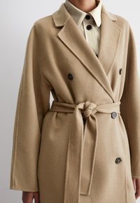 Cappotto doppiopetto in misto lana beige con cintura, grandi bottoni e maniche ampie; texture liscia e design sartoriale.