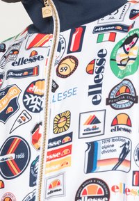 Färgglad vit zip-up jacka med olika logotyper och märken i röd, blå, gul och grön. Gulddragkedja och ribbad marinblå krage.