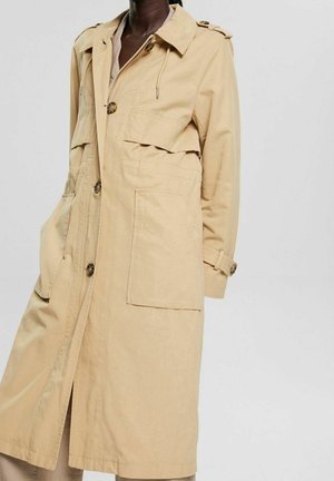 Personne à la peau foncée portant un long trench-coat beige avec de grandes poches avant et des boutons, debout sur un fond clair.