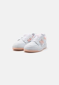 Baskets blanches avec texture perforée, accents roses sur les côtés et le talon, design à lacets, et semelles en caoutchouc. Le modèle présente une forme basse.