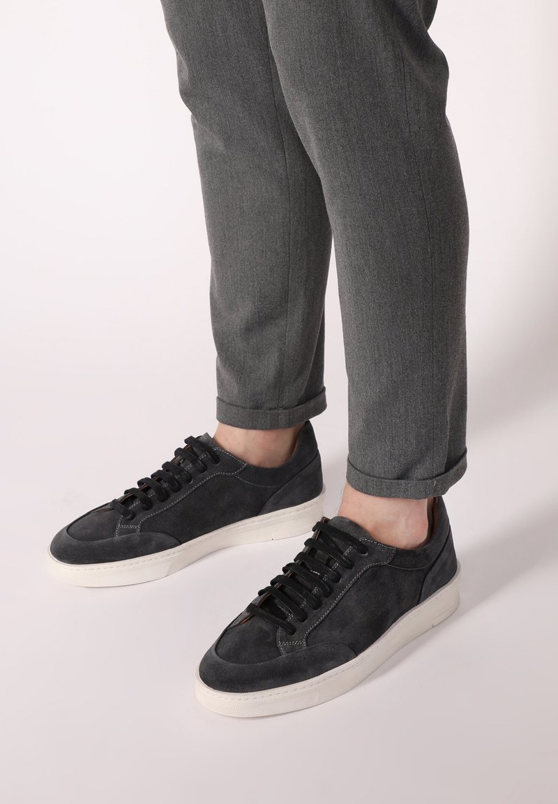 Šedé semišové tenisky s bílými gumovými podrážkami a šňůrkami, se zaoblenou špičkou a minimalistickým designem, párované s úzkými šedými kalhotami.