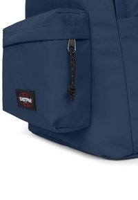 Mochila de tela azul con un bolsillo frontal, detalles de cremallera con un tirador de cordón y un parche de logo negro de Eastpak. Textura suave y forma rectangular.