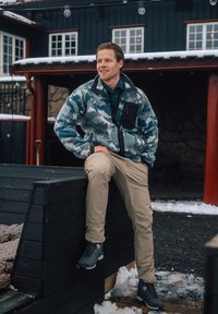 Chaqueta de forro polar para hombre en patrón de camuflaje azul con bolsillo negro, pantalones beige y botas grises. Escenario al aire libre con nieve y estructura de madera.