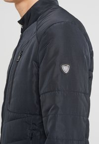 Czarna pikowana kurtka z wysokim kołnierzem, przednią kieszenią zapinaną na zamek oraz naszywką z logo na lewym rękawie. Materiał wydaje się gładki i ocieplany.