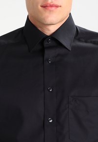 Black button-up shirt with a pointed collar, smooth texture, six buttons, and a single chest pocket featuring a small square accent.