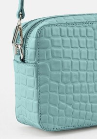 Türkise Tasche mit Krokodilprägung, silbernem Reißverschluss und Beschlägen, ausgestattet mit einer Seitenasche und glatter Oberfläche. Kompakte rechteckige Form.