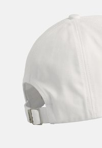 White baseball cap with a textured fabric, curved brim, and adjustable strap at the back. Features a metallic clasp with brand logo.
