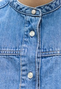 Μπλε τζιν πουκάμισο με στρογγυλεμένο γιακά, δύο κουμπιά στον λαιμό και αντιθετική ραφή κατά μήκος του μπροστινού μέρους και της περιοχής της τσέπης.