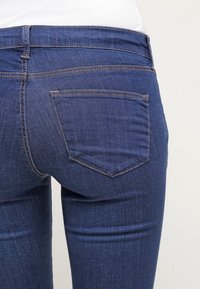 Bakifrån av en person som bär åtsittande blå denimjeans med synlig bakficka och bruna sömmar, tillsammans med en instoppad vit skjorta.