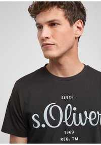 Zwarte katoenen T-shirt met grote witte grafische tekst "s.Oliver" en "Sinds 1969" met een ronde hals en korte mouwen.