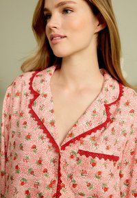 Cămiză de pijama roșie, cu flori, pe un fundal alb, având dantelă pe guler și pe buzunar, cu o croială lejeră și confortabilă.