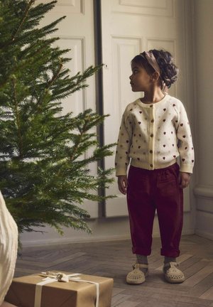 Enfant portant un cardigan crème avec des motifs de cœurs rouges, un pantalon bordeaux et des chaussures en crochet beiges, se tenant près d'un arbre de Noël et d'un cadeau.