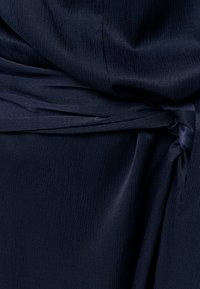 Tamnoplava tkanina s diskretnim teksturiranim uzorkom vezana u labavi čvor, stvarajući nabore i mekane sjene.