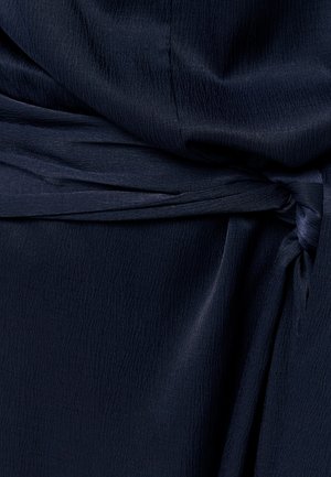 Tamnoplava tkanina s diskretnim teksturiranim uzorkom vezana u labavi čvor, stvarajući nabore i mekane sjene.