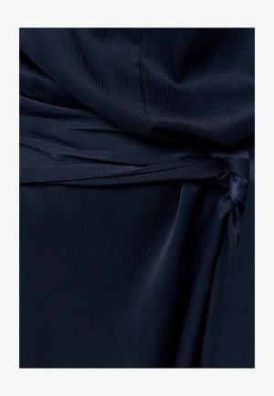 Tela de azul marino oscuro con un patrón texturizado sutil anudada en un nudo suelto, creando pliegues y sombras suaves.