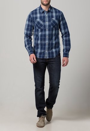 Camicia a quadri blu navy con dettagli azzurri, due tasche sul petto, maniche lunghe e colletto classico, abbinata a jeans scuri e scarpe beige.