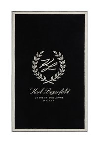 Melns taisnstūra paklājs ar pūkainu tekstūru, ar baltu izšūtā logo un lauvas rotājumu, apmali vieglā pelēkā krāsā.