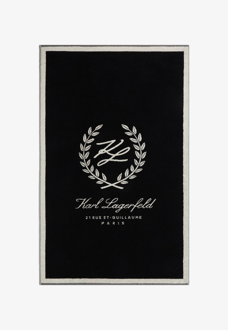 Melns taisnstūra paklājs ar pūkainu tekstūru, ar baltu izšūtā logo un lauvas rotājumu, apmali vieglā pelēkā krāsā.