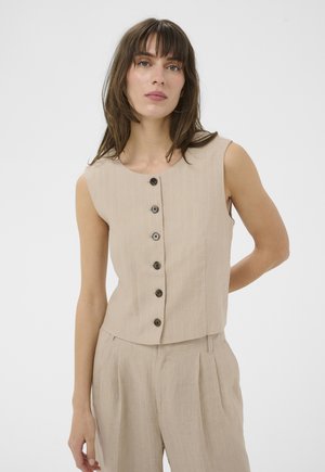 Femme portant un haut beige sans manches à boutons et un pantalon plissé assorti, debout devant un fond blanc uni.