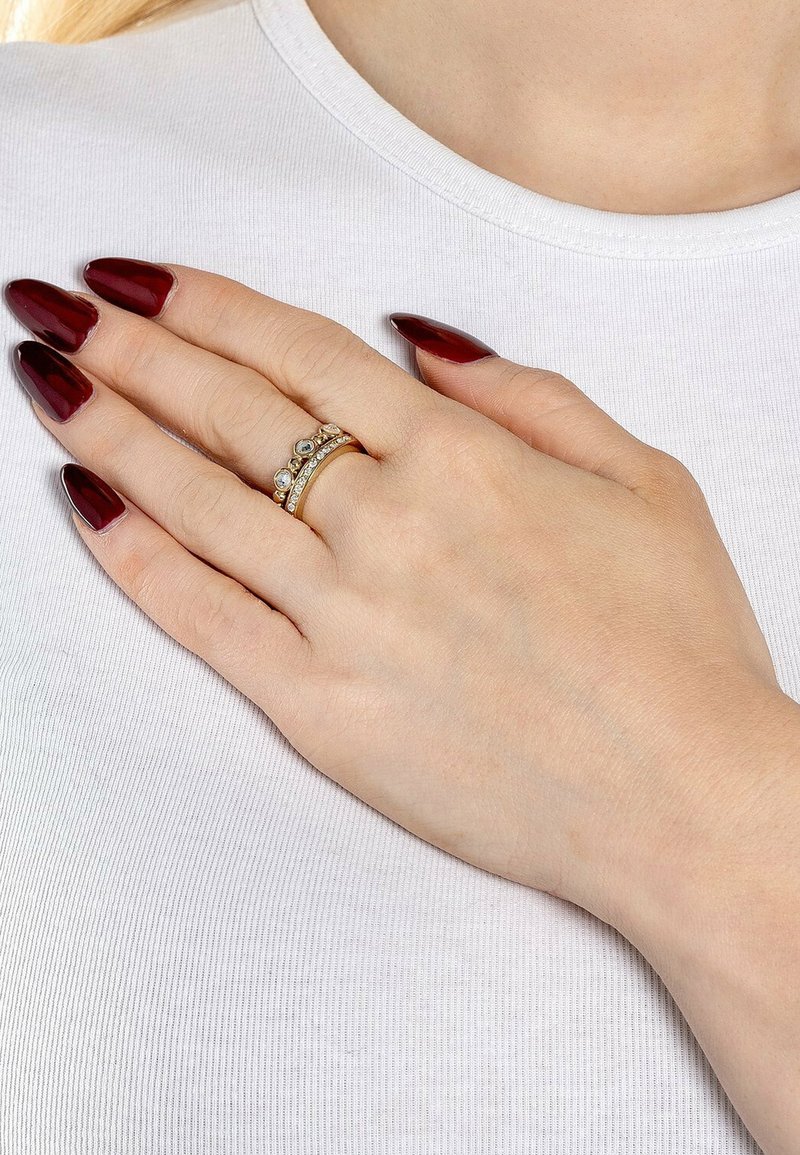 Guldtonad ring med tre band som har klara och mörka stenar, mot en ljus tygbakgrund. Långa, mörk röda naglar synliga.