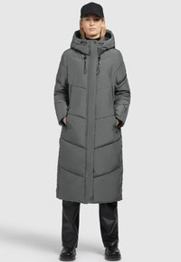 Grijze gewatteerde jas met een capuchon, verticale naden, zijzakken en drukknoopsluitingen aan de zijkant. Draagt met zwarte broek en een pet.