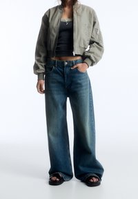 Cazadora bomber gris recortada con puños elásticos, combinada con vaqueros azul descoloridos de pierna ancha. Sandalias negras completan el conjunto.