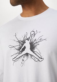 Valkoinen puuvillainen T-paita, jossa on musta siluetti hyppelevästä hahmosta koripallon kanssa, ympäröitynä harmaan sävyisellä räjähdykuviolla.