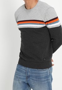 Pull en maille gris et gris foncé avec des rayures horizontales orange, bleu et blanc. Poignets et ourlet côtelés ; écusson du logo près de la taille.