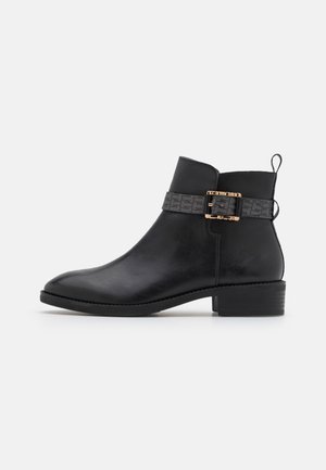 Bottine en cuir noir avec un petit talon bloc, une lanière grise décorative et une boucle dorée sur fond blanc.