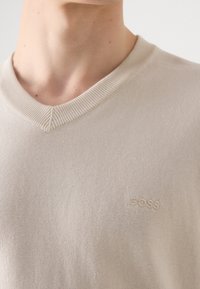 Gros plan sur une personne portant un pull beige à col en V avec un petit logo brodé "BOSS" sur la poitrine.