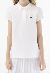Valkoinen polo-paita, joka on valmistettu teksturoidusta puuvillasta. Paidassa on laineileva kaulus, lyhyet puffihihat ja vasemmassa rinnassa vihreä Lacoste-logo.