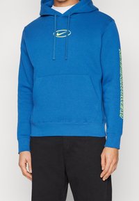 Osoba v modré mikině Nike s neónově žlutým logem a textem na rukávu, v kombinaci s černými kalhotami, na jednolitém pozadí.