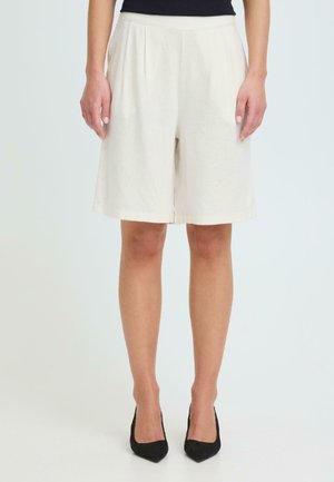 Person iført kremfargede shorts til knelengde og svarte spisse hæler, stående mot en ensfarget lys bakgrunn.