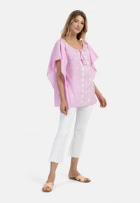 Tunika di cotone rosa con maniche ampie, caratterizzata da motivi floreali ricamati e una scollatura con cordoncino. Abbinata a pantaloni bianchi a 3/4.