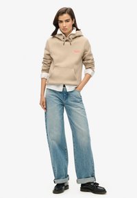 Felpa beige con cappuccio, coulisse, tasca anteriore e logo rosso. Abbinata a jeans azzurri chiari, larghi e scarpe nere.