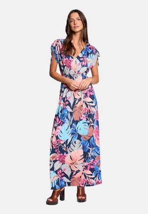 Femme portant une robe maxi fleurie colorée à manches courtes et des sandales compensées marron, debout devant un fond uni.