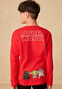 Červená košile s dlouhým rukávem, která na zadní straně nese bílé logo "STAR WARS" a v barvě má vytištěné karikatury postav Darth Vader, Yoda a R2-D2.