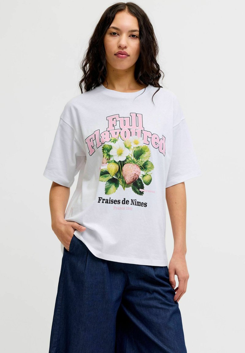 Νεαρή γυναίκα που φοράει λευκό φαρδύ T-shirt με ροζ κείμενο "Full Flavoured" και σχέδιο από φράουλες με λουλούδια, συνδυασμένο με σκούρο μπλε παντελόνι.