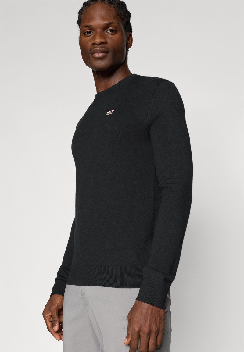 Maglione nero lavorato a maglia con scollo a girocollo, polsini e orlo a costine, caratterizzato da un piccolo logo sul petto. Tessuto liscio e vestibilità sagomata.