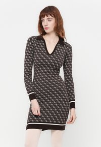 Donna che indossa un abito marrone a maniche lunghe e aderente, con motivi geometrici bianchi e bordi bianchi sui polsini e sull'orlo, su uno sfondo bianco uniforme.