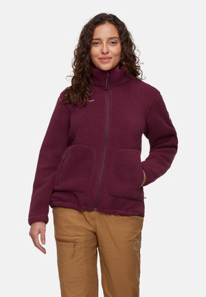 Veste en polaire bordeau avec un col élevé, deux poches latérales, fermeture éclair et tissu texturé. Portée par-dessus un pantalon marron.